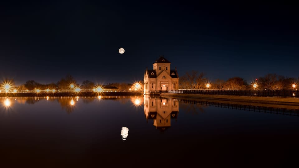 A lit church with a reflection in a calm body of water, surrounded by a fence and trees under a night sky.