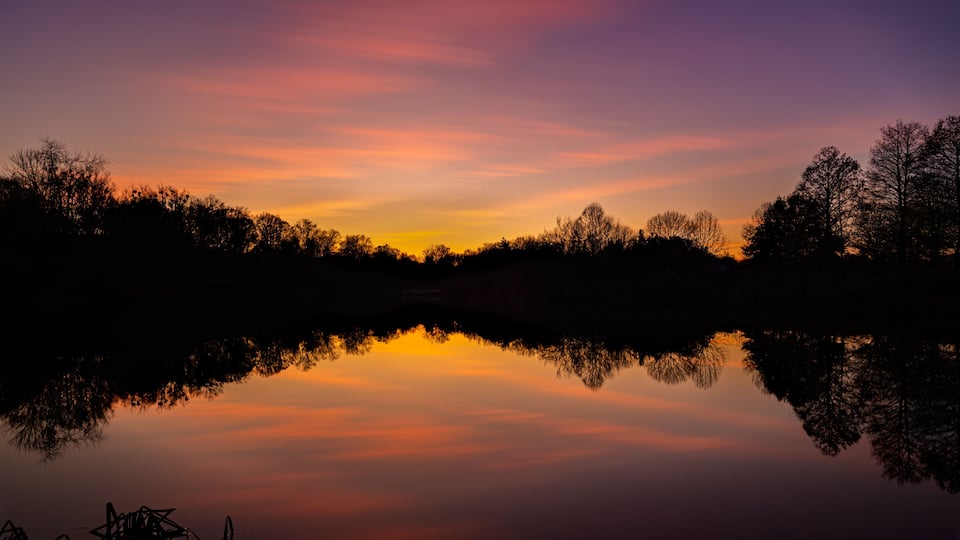 A sunset over a calm lake with trees silhouetted against the sky.