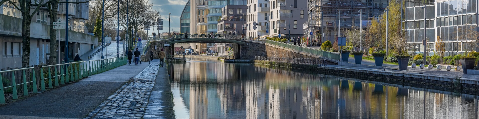 A canal with a stone-paved walkway, a bridge, and buildings with modern architecture.