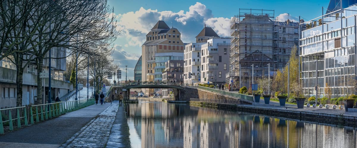 A canal with a stone-paved walkway, a bridge, and buildings with modern architecture.