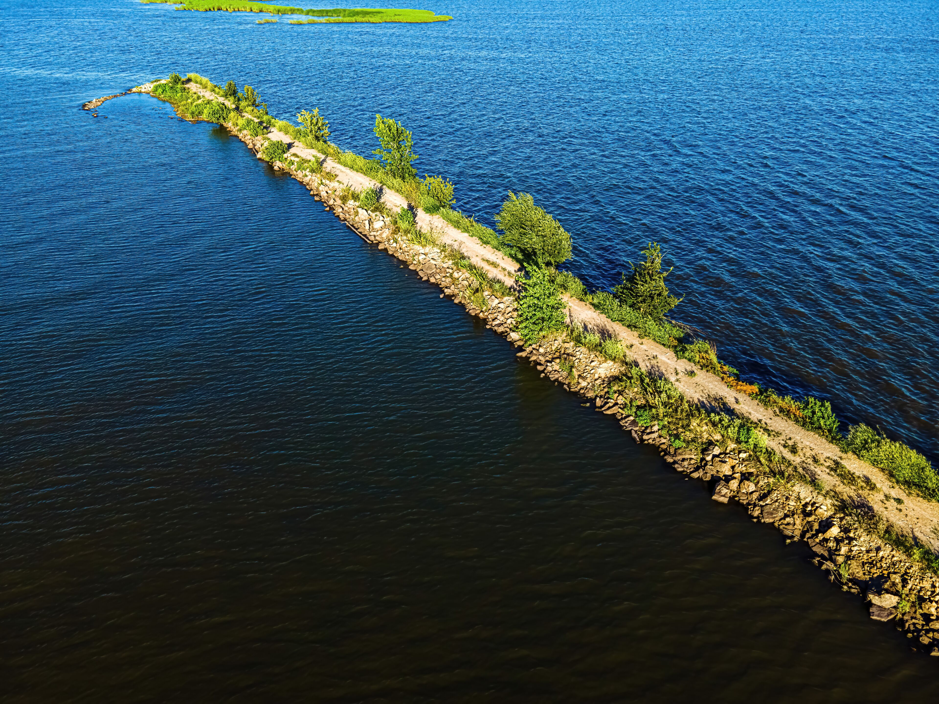 Una isla estrecha y rocosa con un sendero de tierra y vegetación, rodeada de aguas tranquilas.