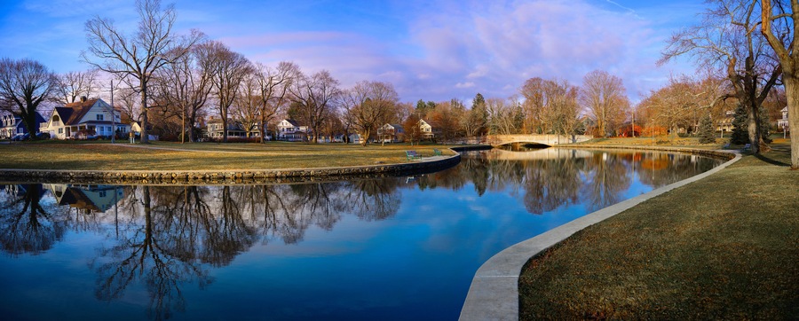 Milford Connecticut City Landscape at the Lower Lagoon Riverwalk Park in winter with curving calm river, green field, bare maple and oak trees, and traditional charming New England houses at sunset