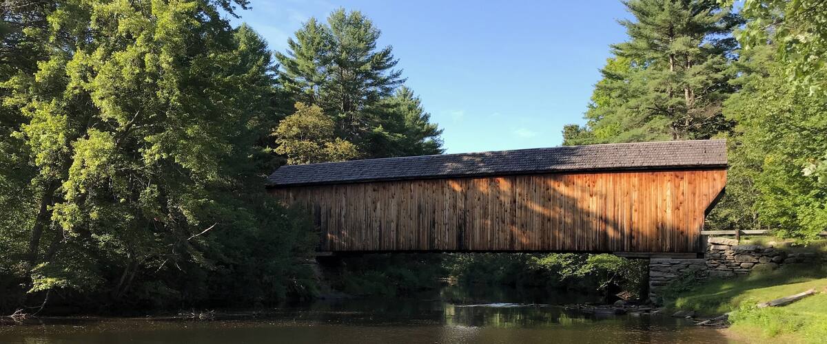 A wooden covered bridge over a calm river, surrounded by lush greenery and trees.