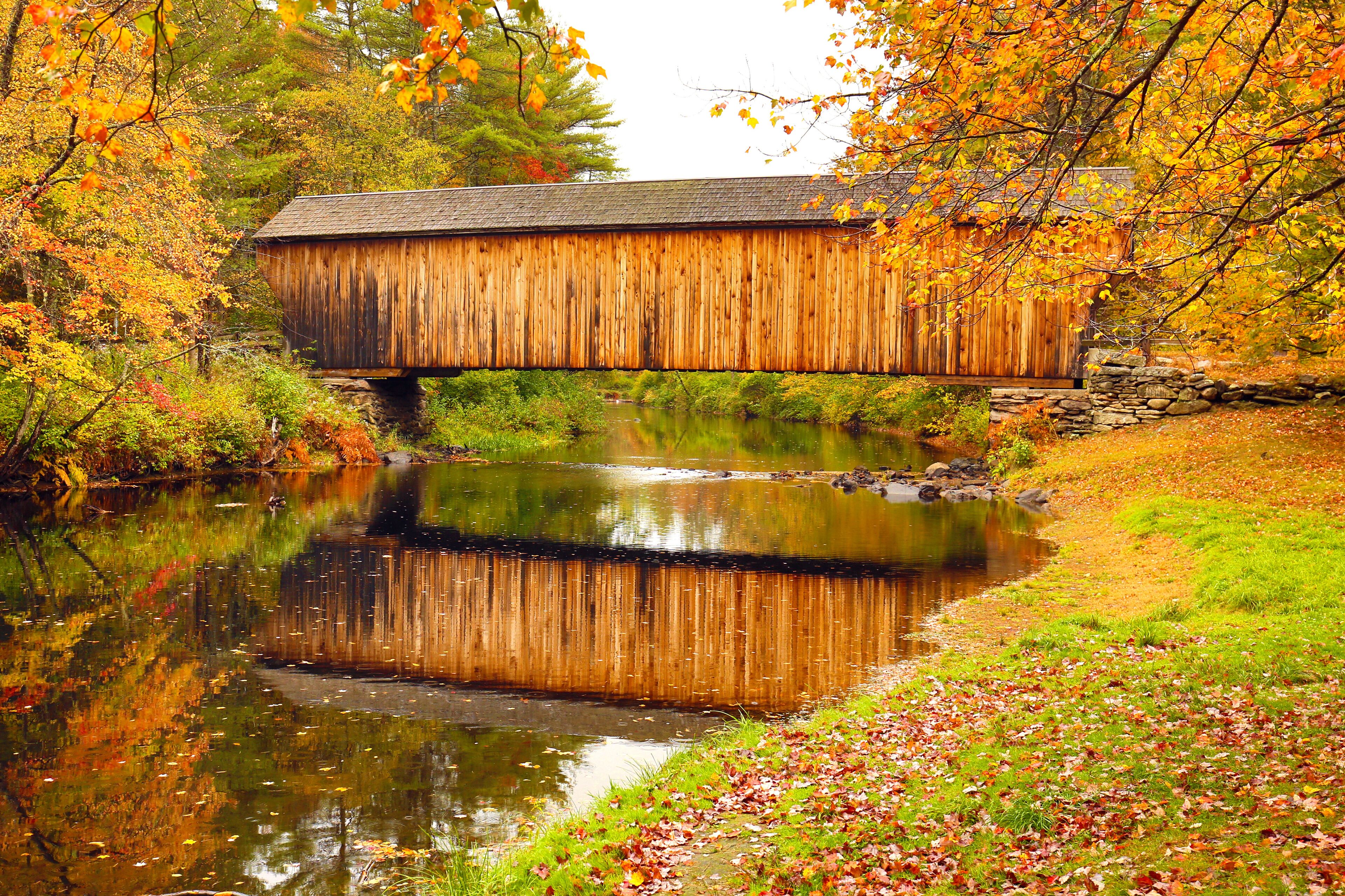 A wooden covered bridge over a calm river surrounded by autumn foliage.