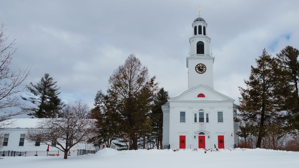 A white church with a clock tower and red doors, surrounded by snow and trees.