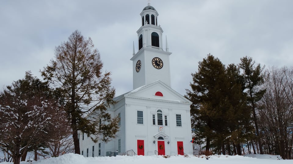A white church with a clock tower, surrounded by snow-covered ground and trees.