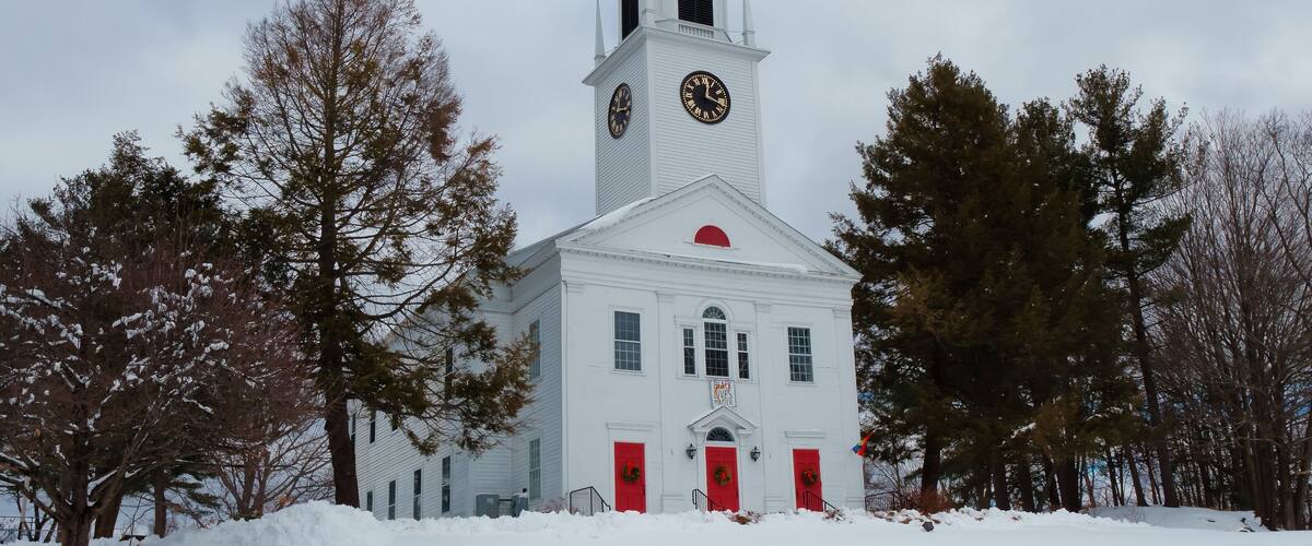 A white church with a clock tower, surrounded by snow-covered ground and trees.