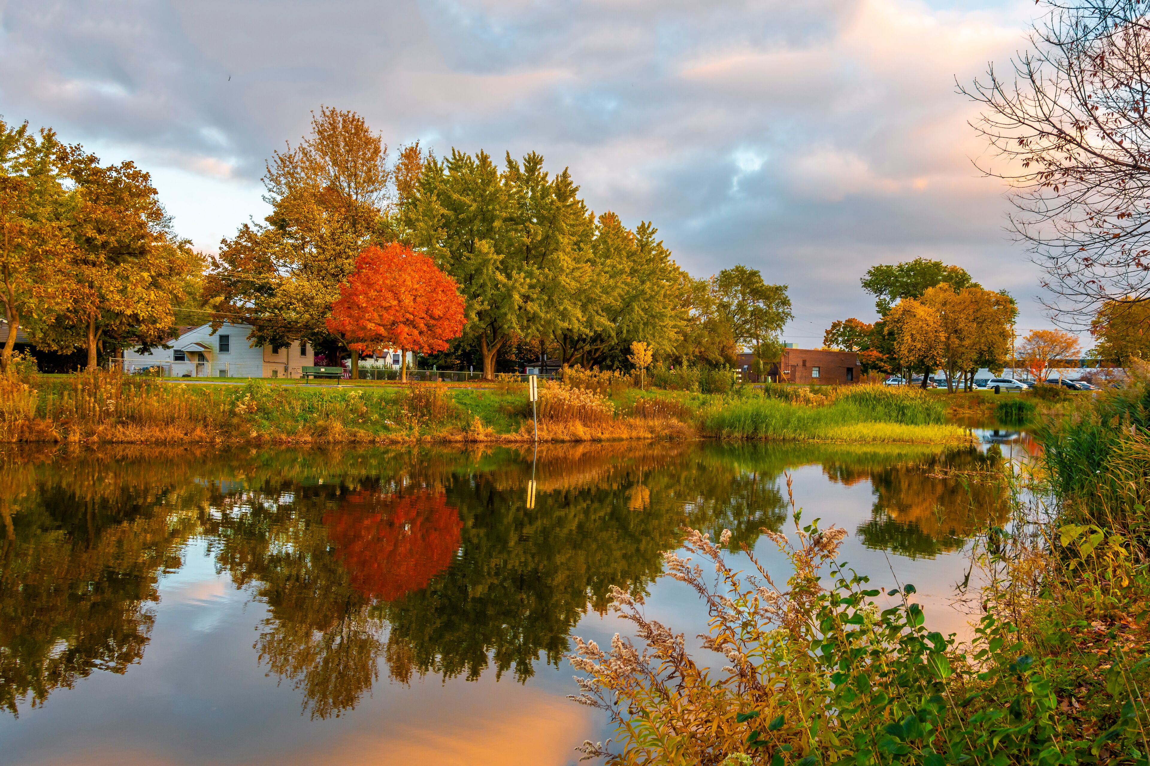 A calm lake with reflections of trees and houses, surrounded by autumn foliage.