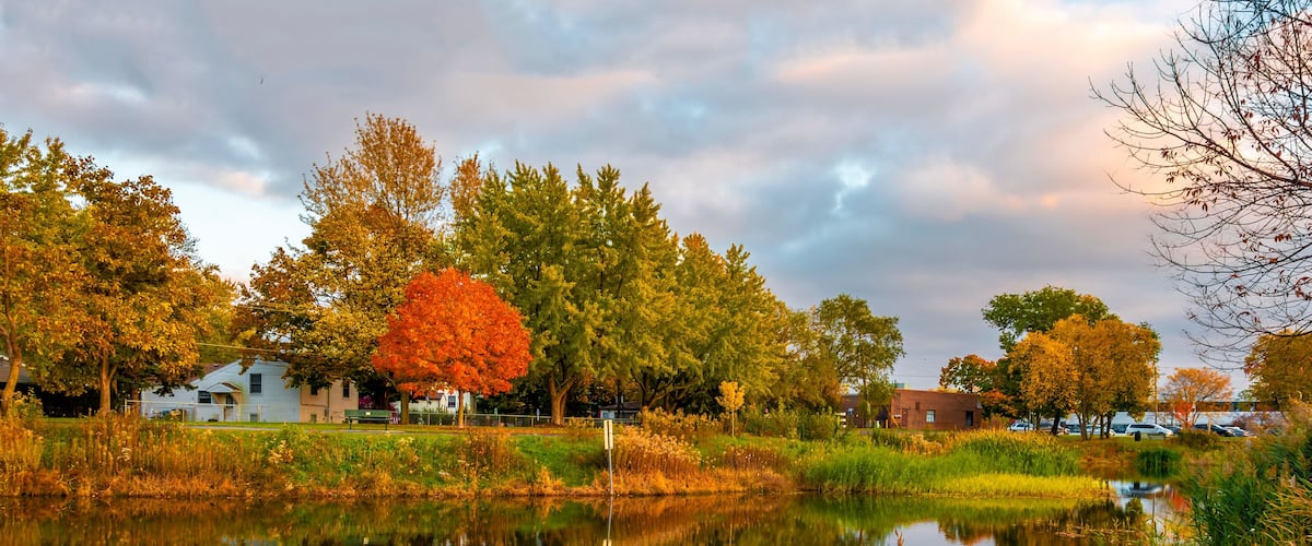 A calm lake with reflections of trees and houses, surrounded by autumn foliage.