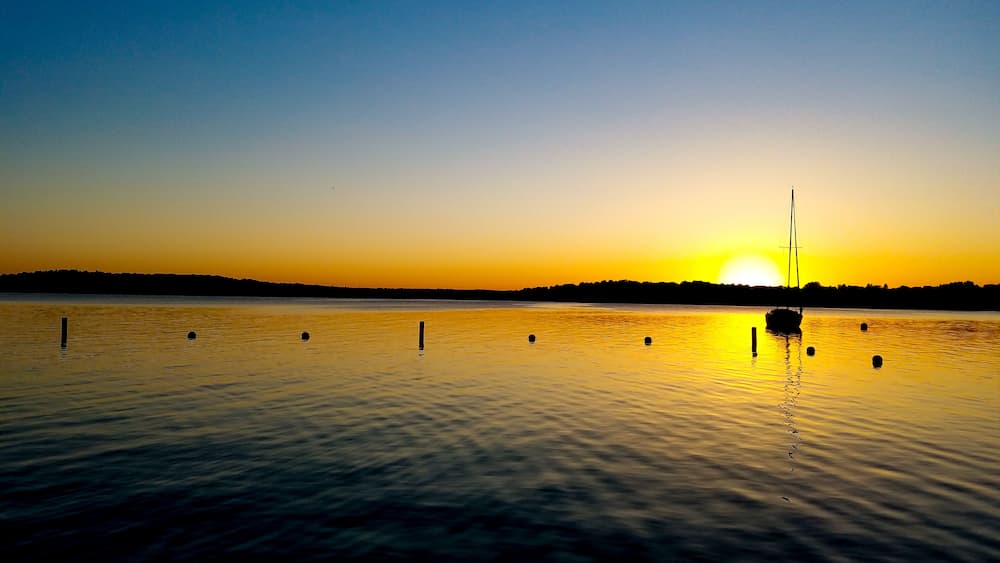 Ein Sonnenuntergang über einem stillen See mit einem Segelboot und Bojen.