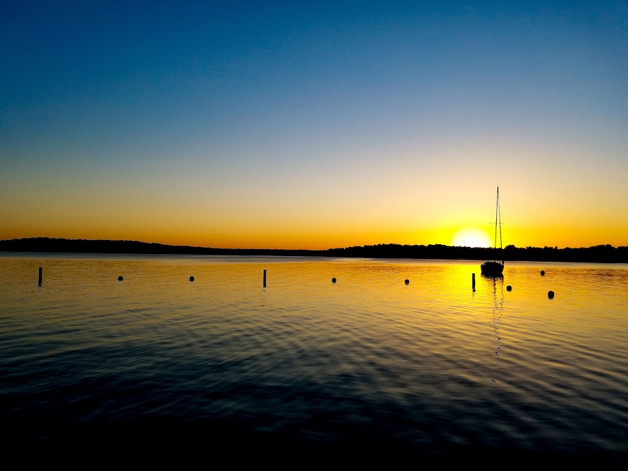 Un coucher de soleil sur un lac paisible, avec un voilier et des bouées flottantes.