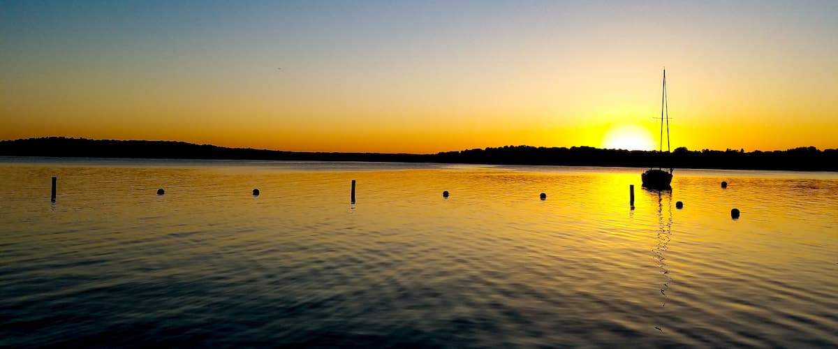 A sunset over a calm lake with a sailboat and floating markers.