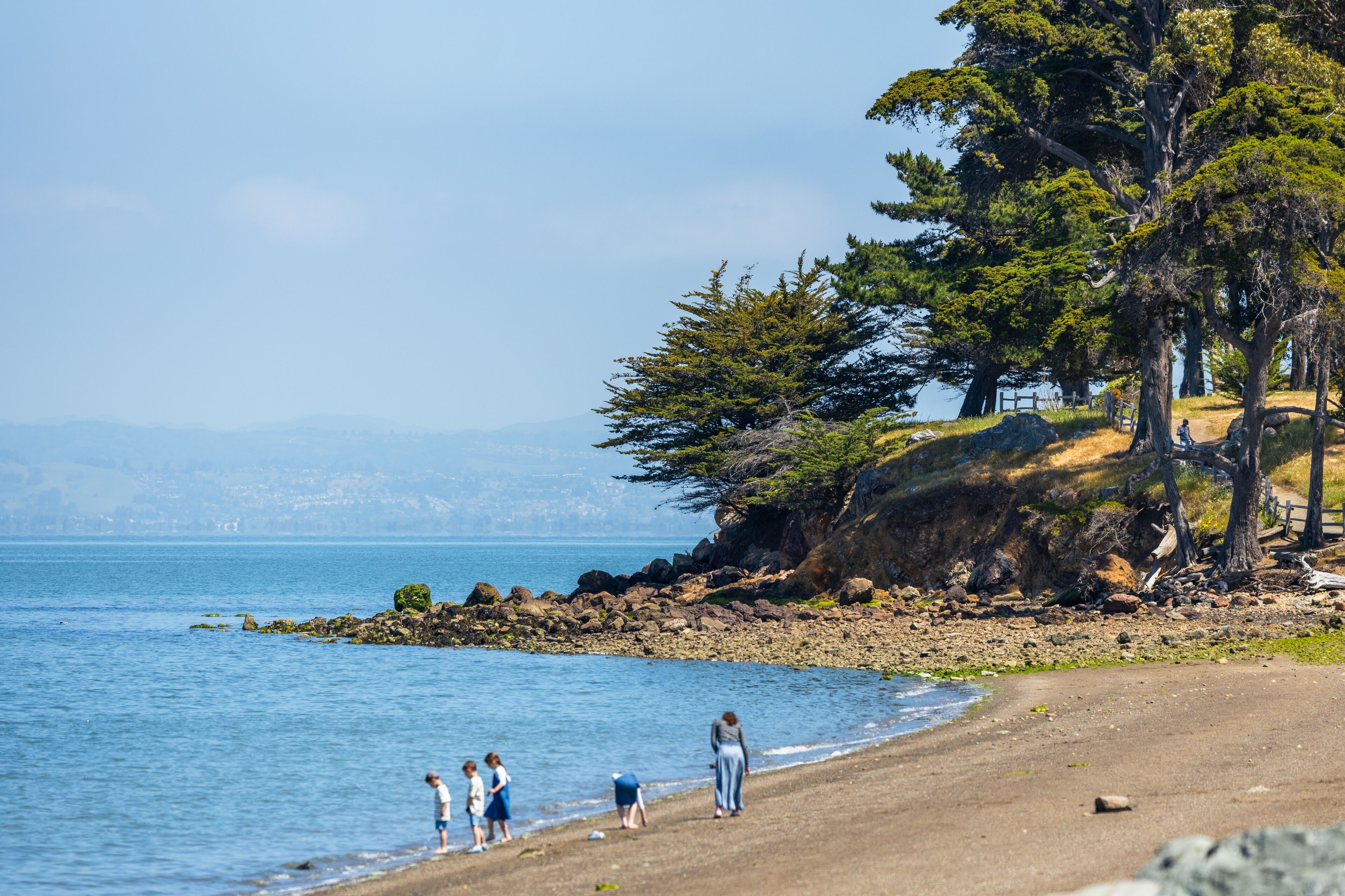 A beach with people walking along the shore, a rocky coastline, and trees in the background.
