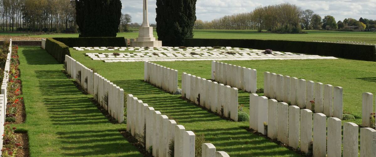 A cemetery with white headstones arranged in rows, a central monument with a cross, and a well-maintained lawn.