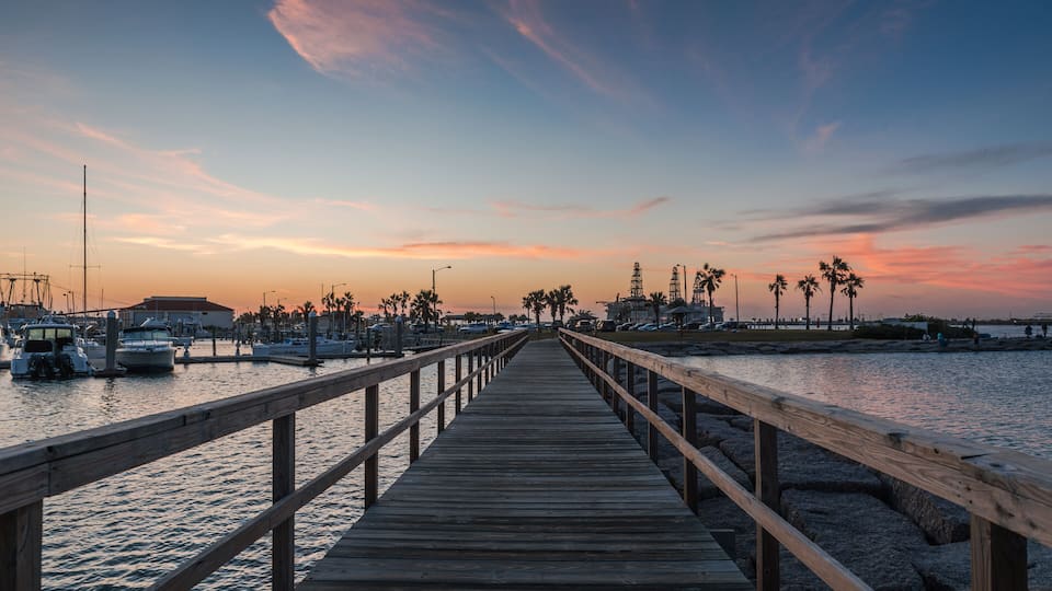 A wooden pier leading to a marina with boats and palm trees under a sunset sky.