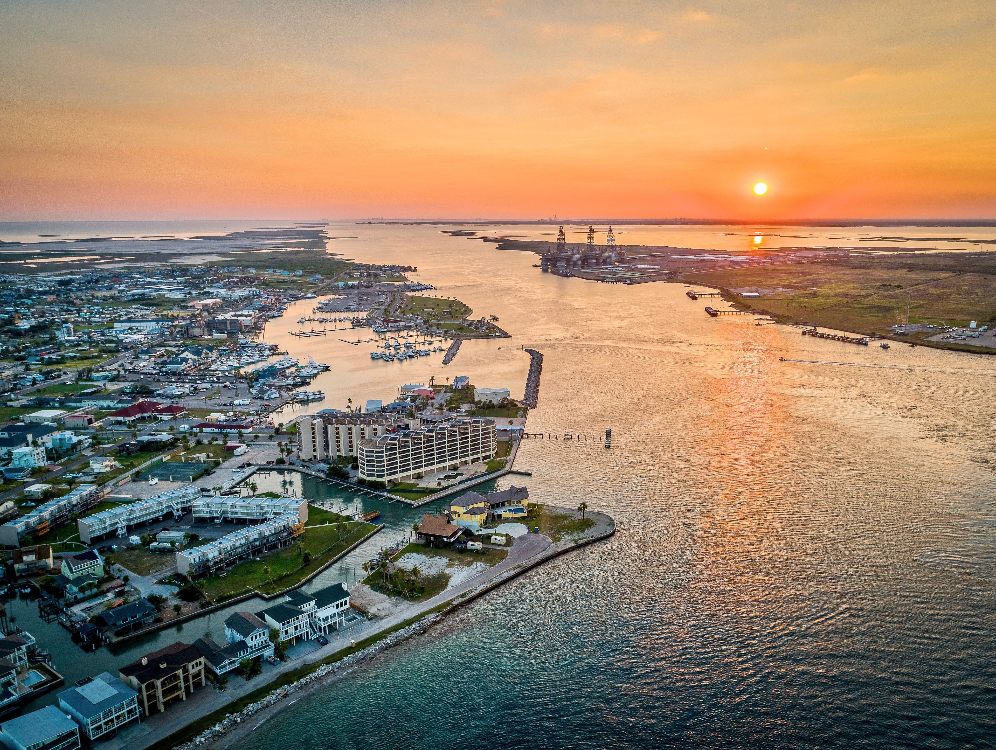 A coastal cityscape at sunset with a marina, residential buildings, and a large hotel.