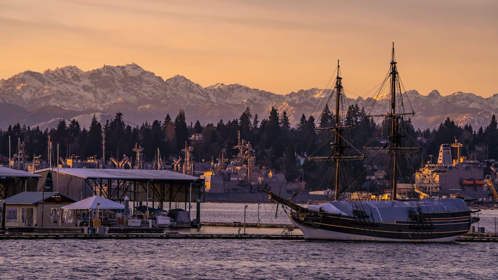 A sailing ship docked at a harbor with mountains in the background.