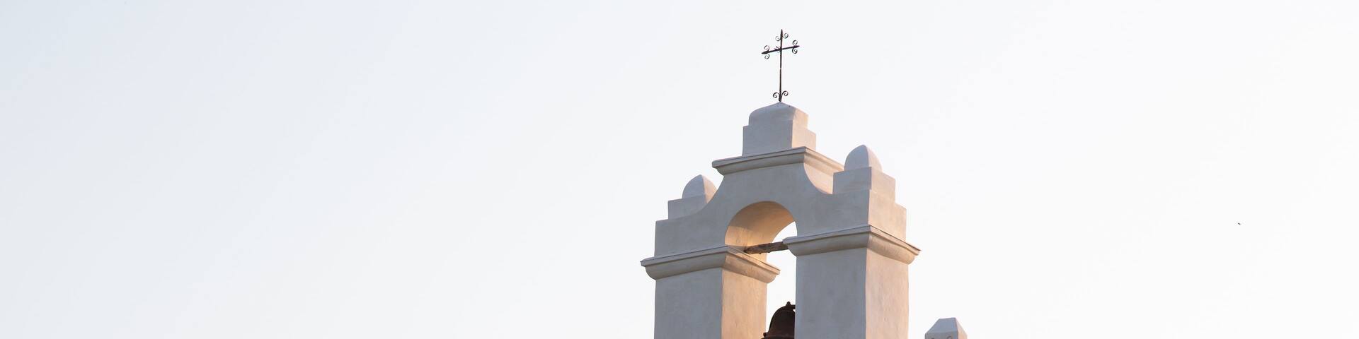 A white bell tower with a cross on top, multiple bells, and arched windows.