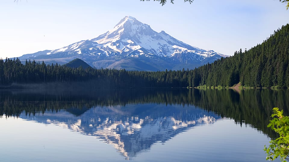 A snow-capped mountain reflected in a calm lake surrounded by dense forests.
