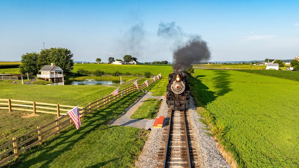A steam locomotive on tracks with American flags, a small house, and open fields.