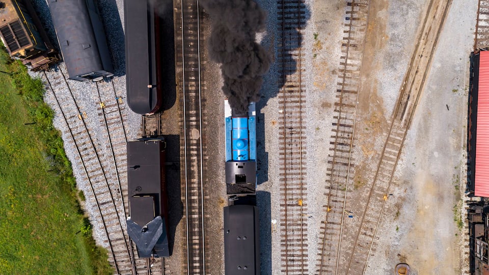 An aerial view of a railway station with multiple tracks, a steam locomotive emitting smoke, and a blue train car.