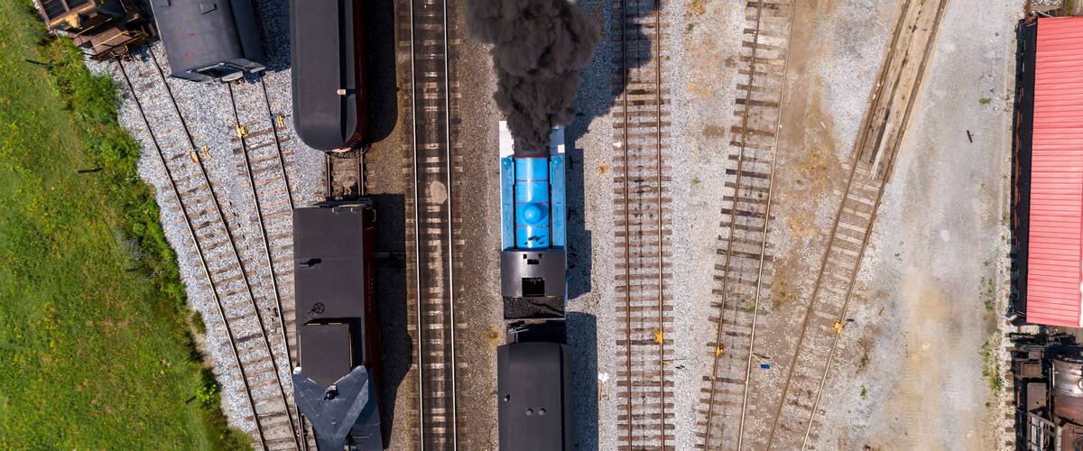 An aerial view of a railway station with multiple tracks, a steam locomotive emitting smoke, and a blue train car.