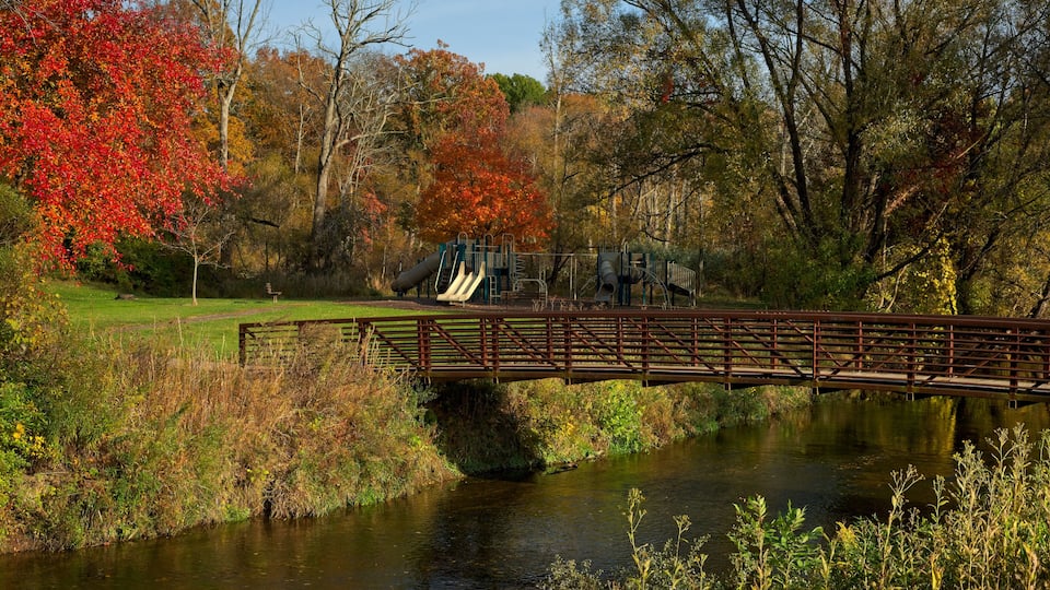 A wooden bridge over a calm river, surrounded by autumn foliage and a playground.