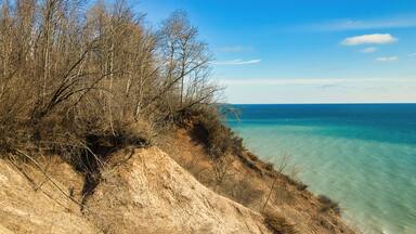 Under a sunny blue sky in early Spring, the view of a peaceful Lake Michigan as seen from atop a sandy, forested bluff at Lion's Den Gorge, near Grafton, WI.