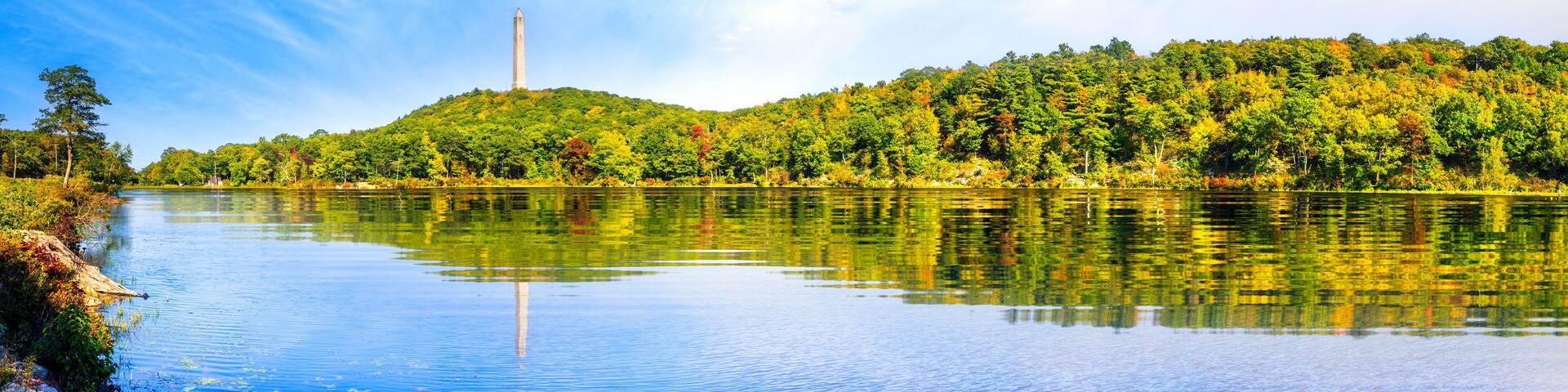 A calm river with a tall monument on a hill, surrounded by dense green forest.