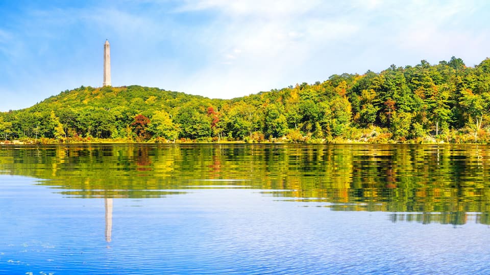 A calm river with a tall monument on a hill, surrounded by dense green forest.