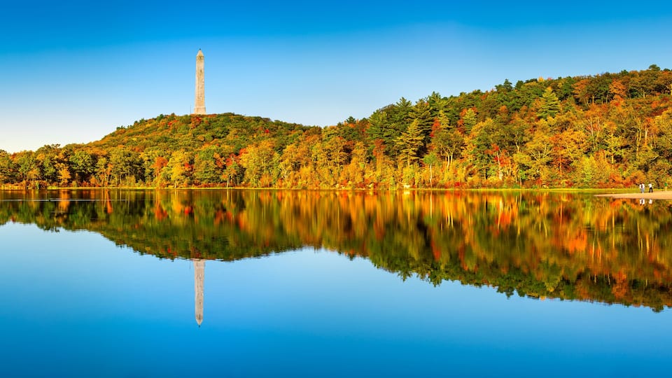 A calm lake with a reflection of a tall monument and a forested hill with autumn foliage.