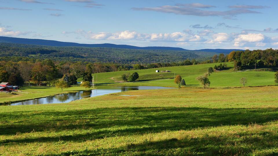 A rural landscape with rolling hills, a pond, and a red barn.