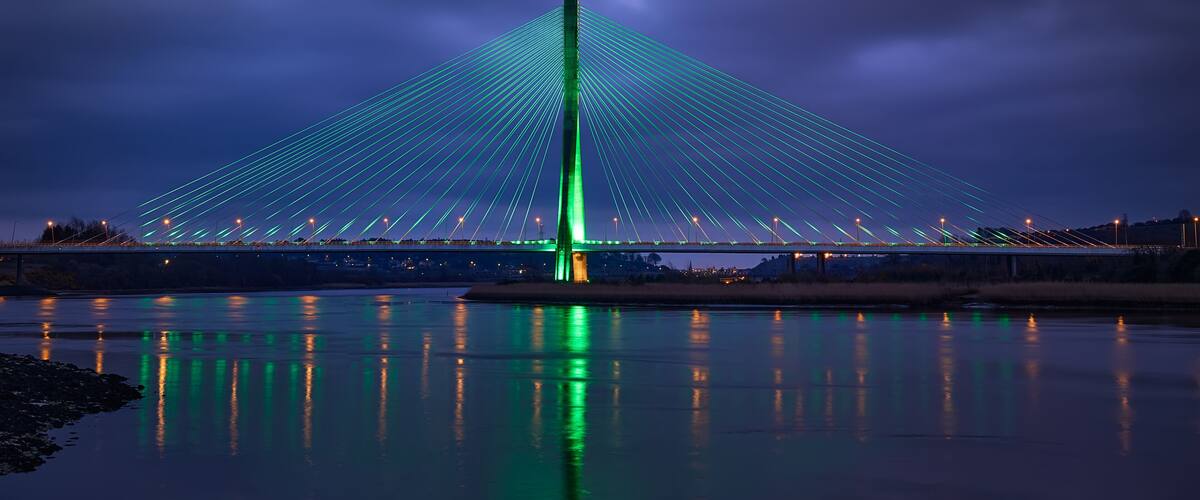 A bridge with a central tower and cables, illuminated at night, spanning a body of water.