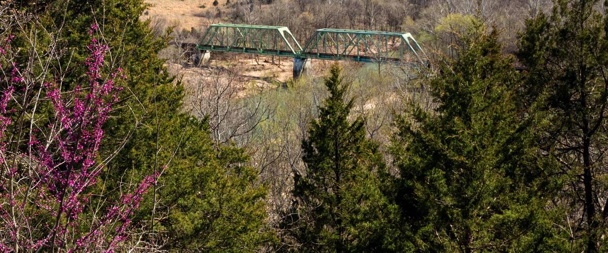 A green metal bridge in a forested area with a backdrop of hills.