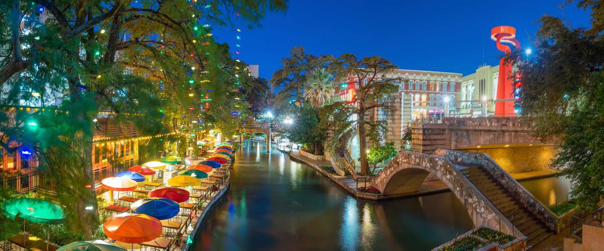 A riverside walkway with colorful umbrellas and a stone bridge.