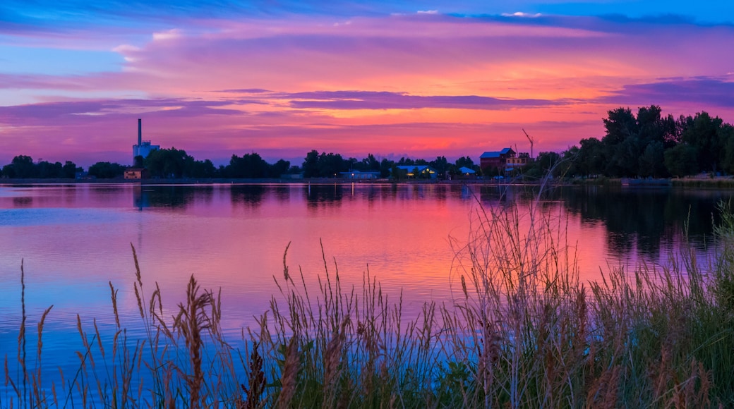 Un atardecer sobre un lago tranquilo con una estructura industrial distante y una hilera de casas a lo largo de la orilla.