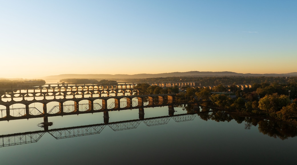 Un puente con múltiples arcos cruzando un río, rodeado de árboles y colinas al fondo.