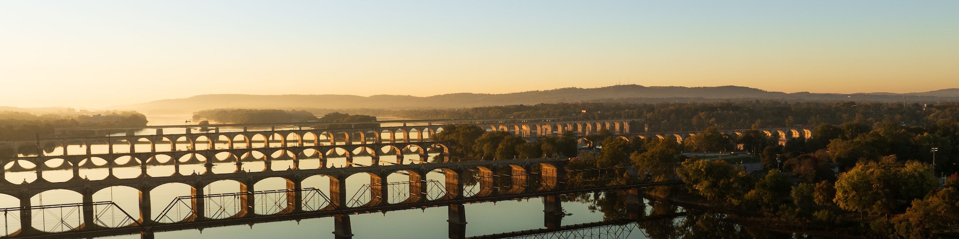 Uma ponte com várias arcos atravessando um rio, cercada por árvores e colinas ao fundo.
