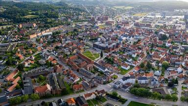 An aerial view of a densely populated town with red-roofed houses, a central square, and a body of water in the background.