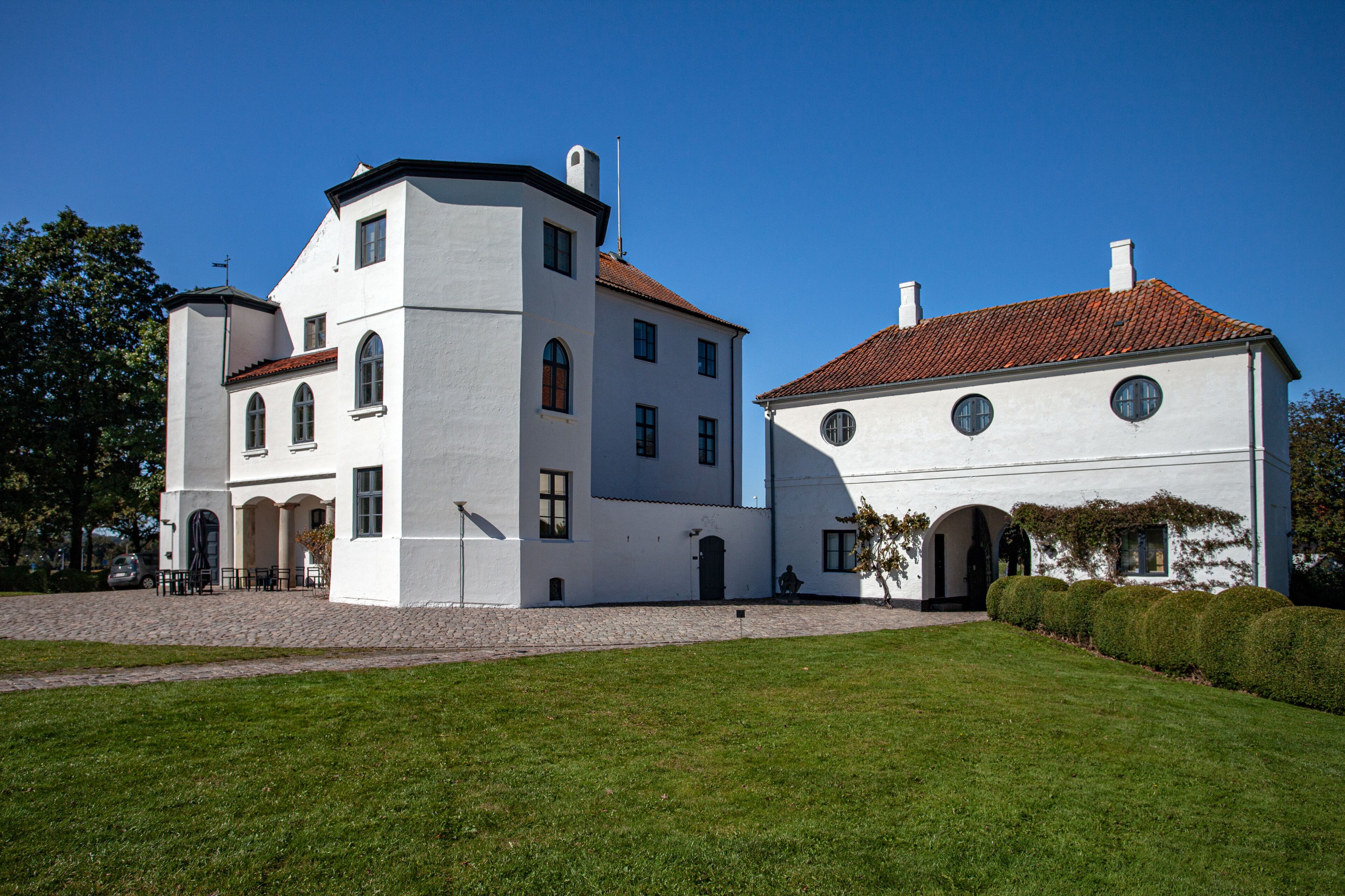 A white building with a red-tiled roof, a cobblestone courtyard, and a well-maintained lawn.