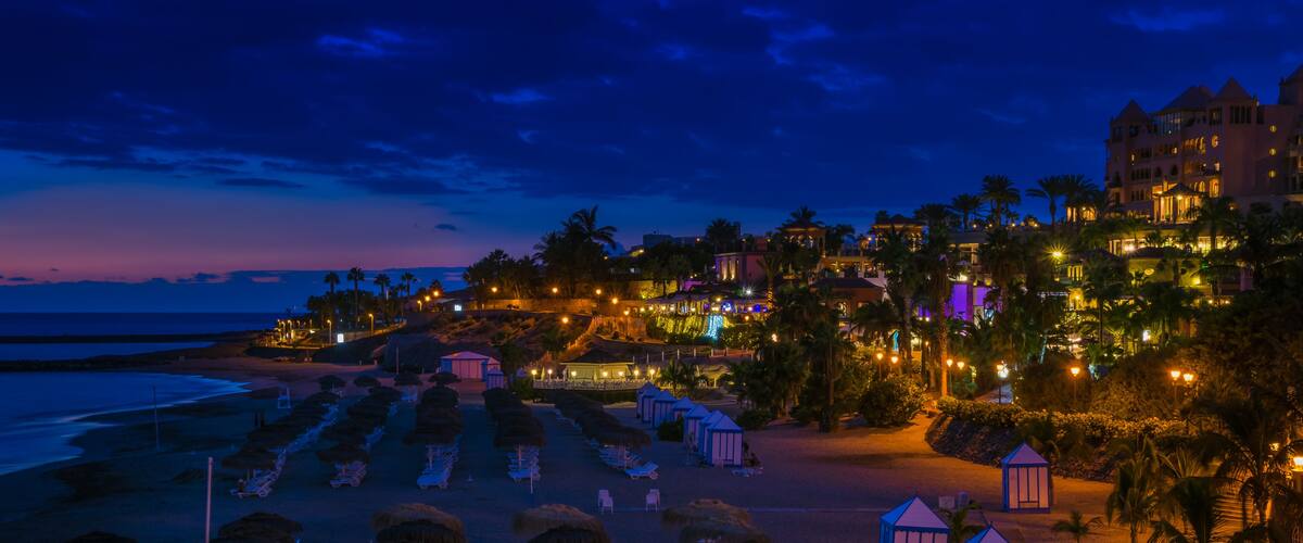 A beach with rows of sun loungers and thatched umbrellas, palm trees, and a brightly lit resort with multiple buildings.