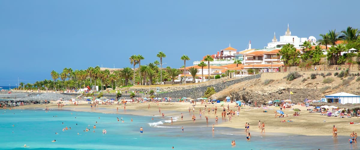 A crowded beach with clear turquoise waters, palm trees, and buildings in the background.