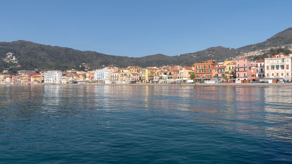 A coastal town with colorful buildings along the waterfront, a clear blue sky, and hills in the background.
