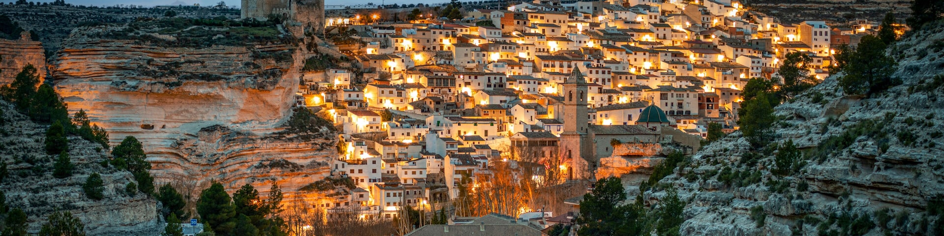 Uma cidade aninhada num vale, com um castelo imponente numa colina, rodeada por terreno rochoso e vegetação densa.