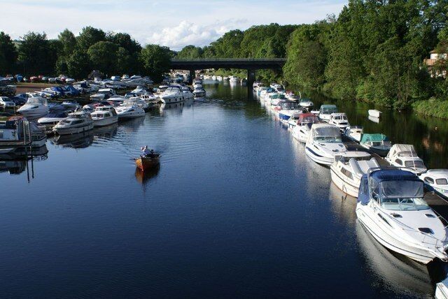 Een jachthaven met tal van boten die langs een rivier liggen, een brug op de achtergrond en bomen langs de rivieroever.
