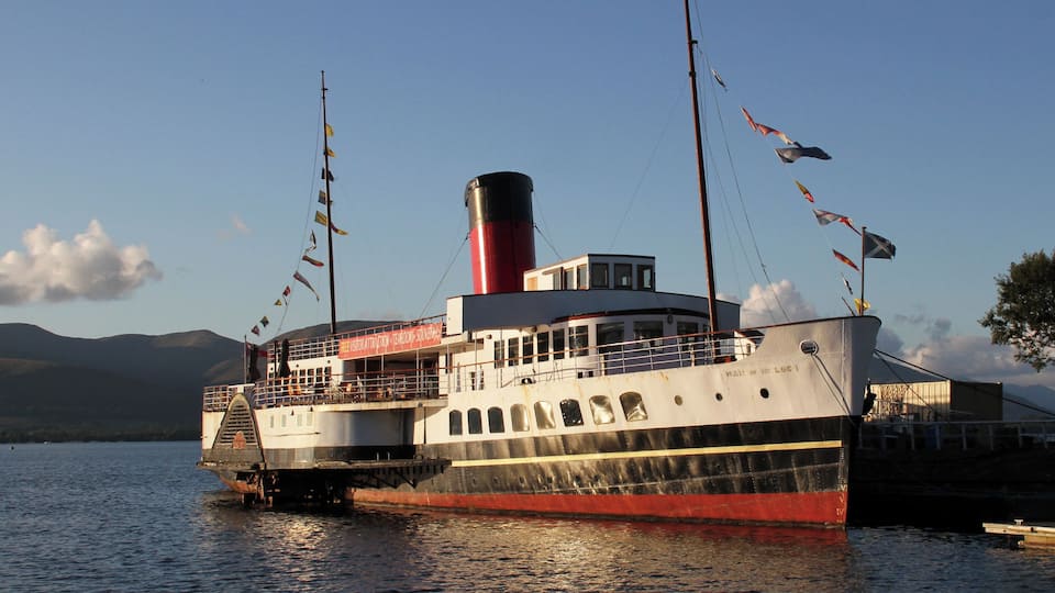 A large, multi-decked steamboat docked at a calm lake with mountains in the background.
