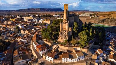 Uma cidade histórica com uma torre de castelo imponente, rodeada por edifícios tradicionais e uma paisagem montanhosa.