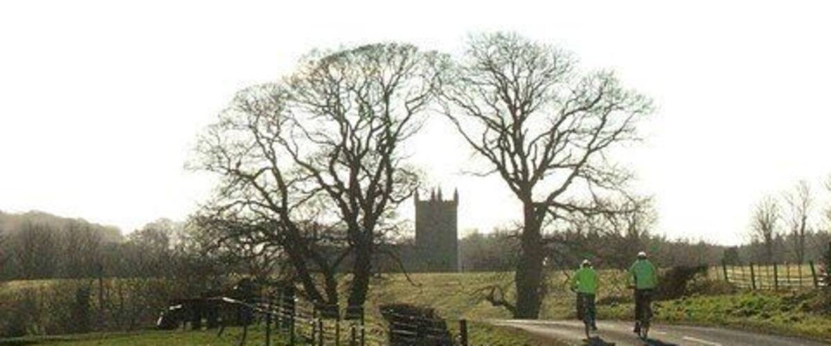 A rural road with cyclists, a stone wall, and a distant church tower.