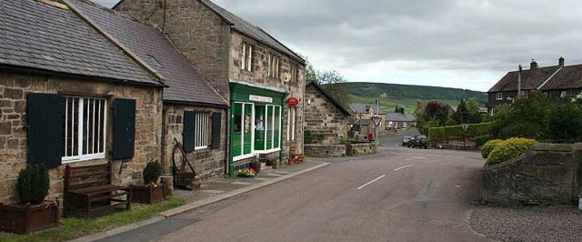 A quiet village street with stone buildings, a green shop, and a hill in the background.