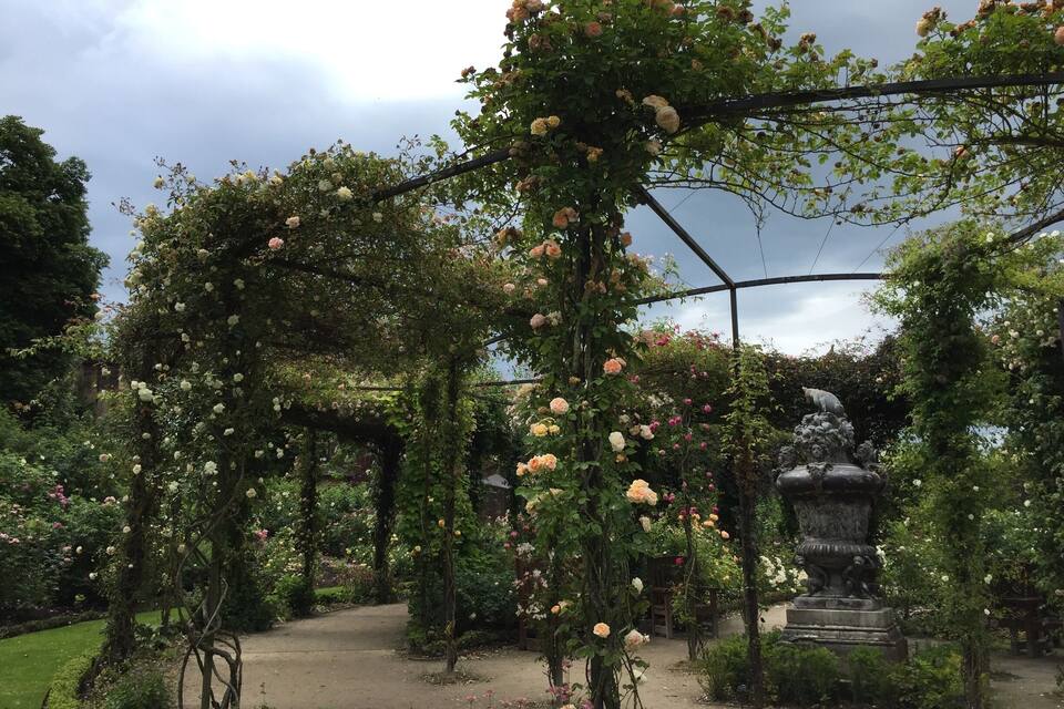 Een tuin met rozenbogen en een standbeeld.